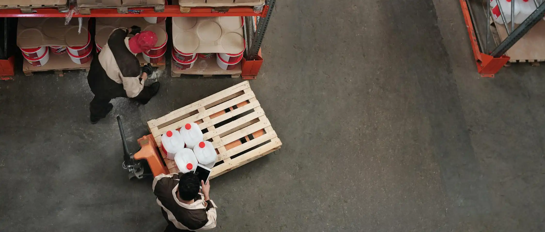 Two workers in a manufacturing warehouse are moving white plastic containers with orange lids on a wooden pallet using a pallet jack. There are shelves with protective products in the background, and the warehouse floor is gray.