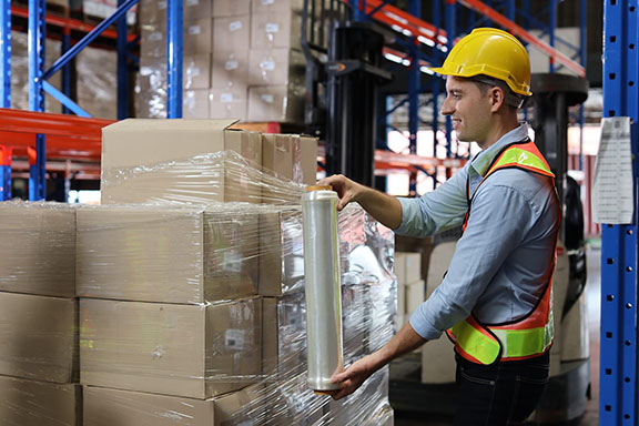 Caucasian warehouse worker man with hardhat and reflective jackets wrapping boxes in stretch film parcel on pallet while control stock and inventory in retail warehouse logistics distribution centre