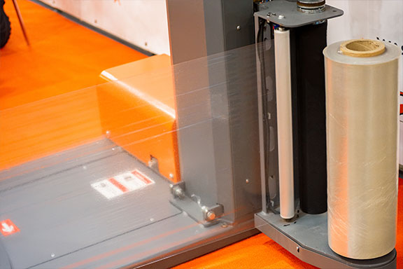 A stretch wrap machine with a roll of pallet wrap film, set against an orange floor.