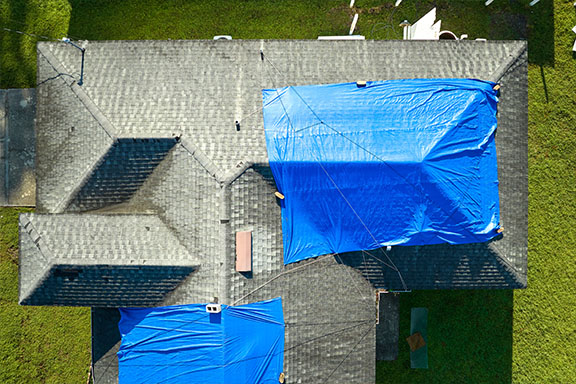 Aerial view of a house with a gray shingle roof, partially covered with blue tarps. Tarps are secured on both sides, indicating roof repairs or protection. The house is surrounded by green grass.