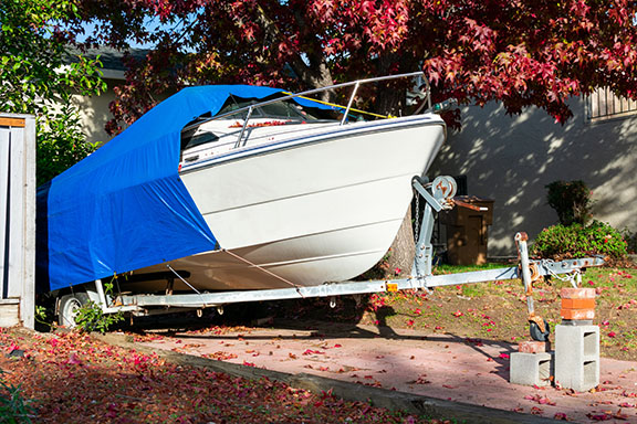 A white boat, shielded by a blue waterproof tarpaulin, is parked on a trailer in the driveway. Autumn trees with vibrant red leaves encircle the area, their fallen foliage scattered across the ground.